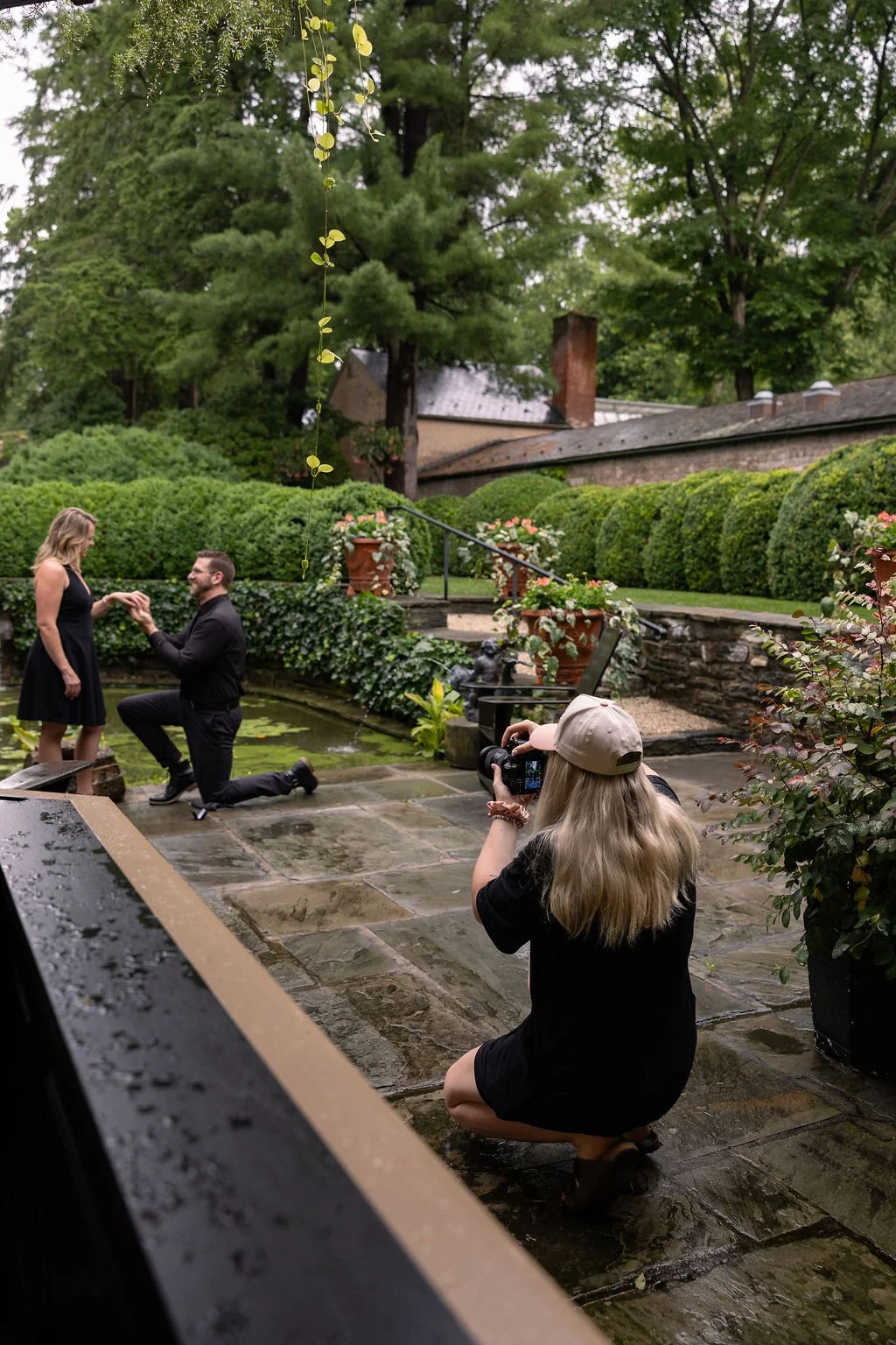 A woman with long blonde hair and a baseball cap is kneeling on wet stone pavement, taking a photo of a romantic proposal scene where a man is kneeling on one knee proposing to a woman in an outdoor garden setting with lush greenery and potted flower