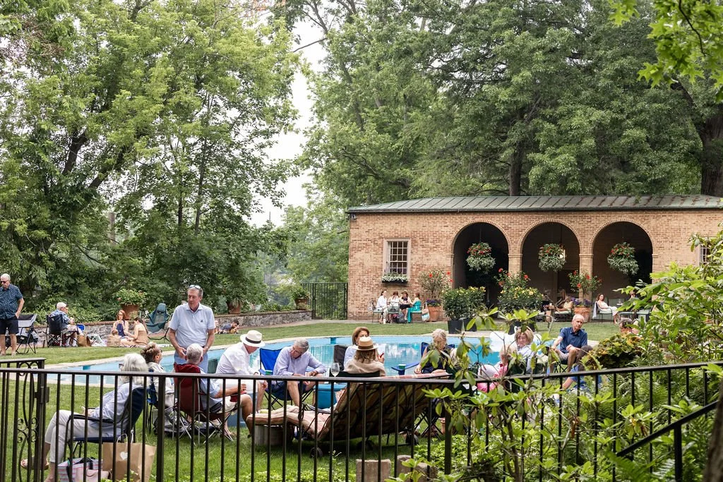 People gathered around a swimming pool in a backyard with lush green trees and a brick building with arches and hanging flower baskets in the background.