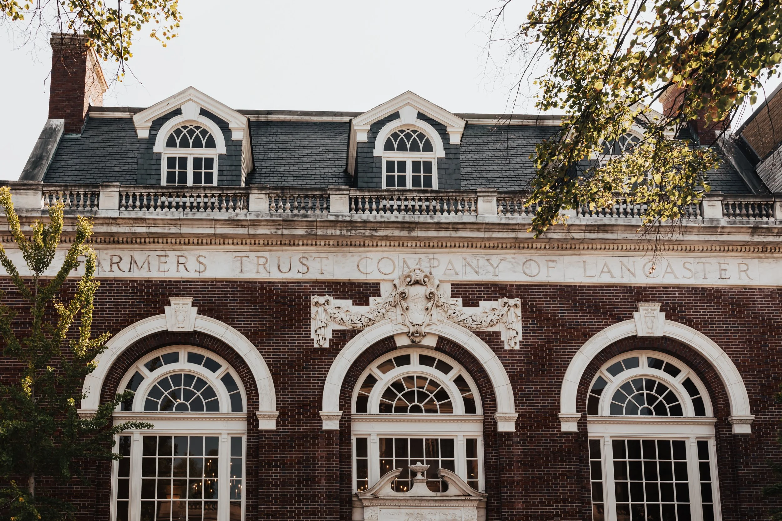 Historic brick building with white decorative trim and arched windows, inscription reading 'Farmers Trust Company of Lancaster' on facade, and a decorative stone emblem above the entrance.