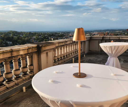 Round table with a white tablecloth and a gold lamp on top, situated on a balcony with city and sky view in the background.
