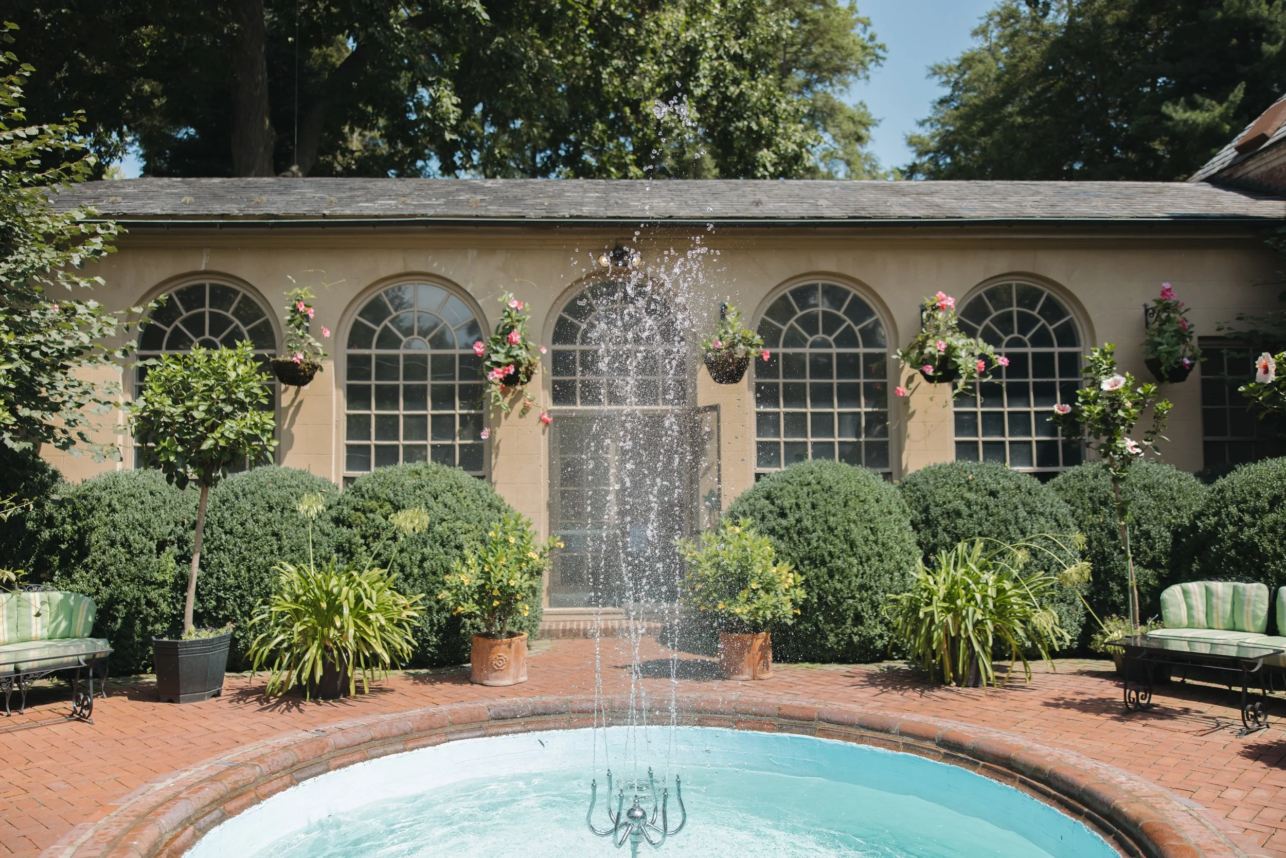 A daytime view of a backyard with a fountain in the foreground, surrounded by potted plants and green bushes, and a house with large arched windows decorated with hanging flower baskets in the background.