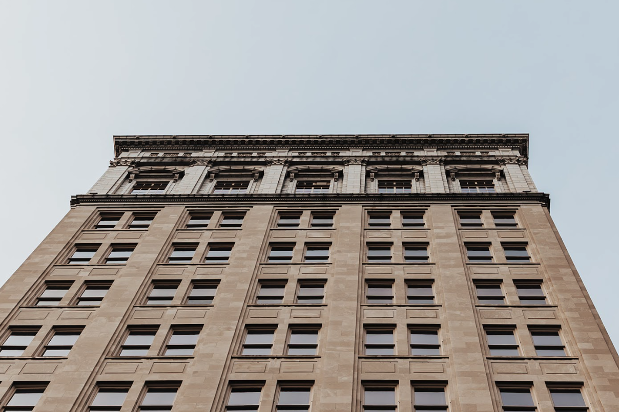 Looking up at a tall, historic office building with detailed stonework, large windows, and decorative architectural elements at the top against a pale, overcast sky.