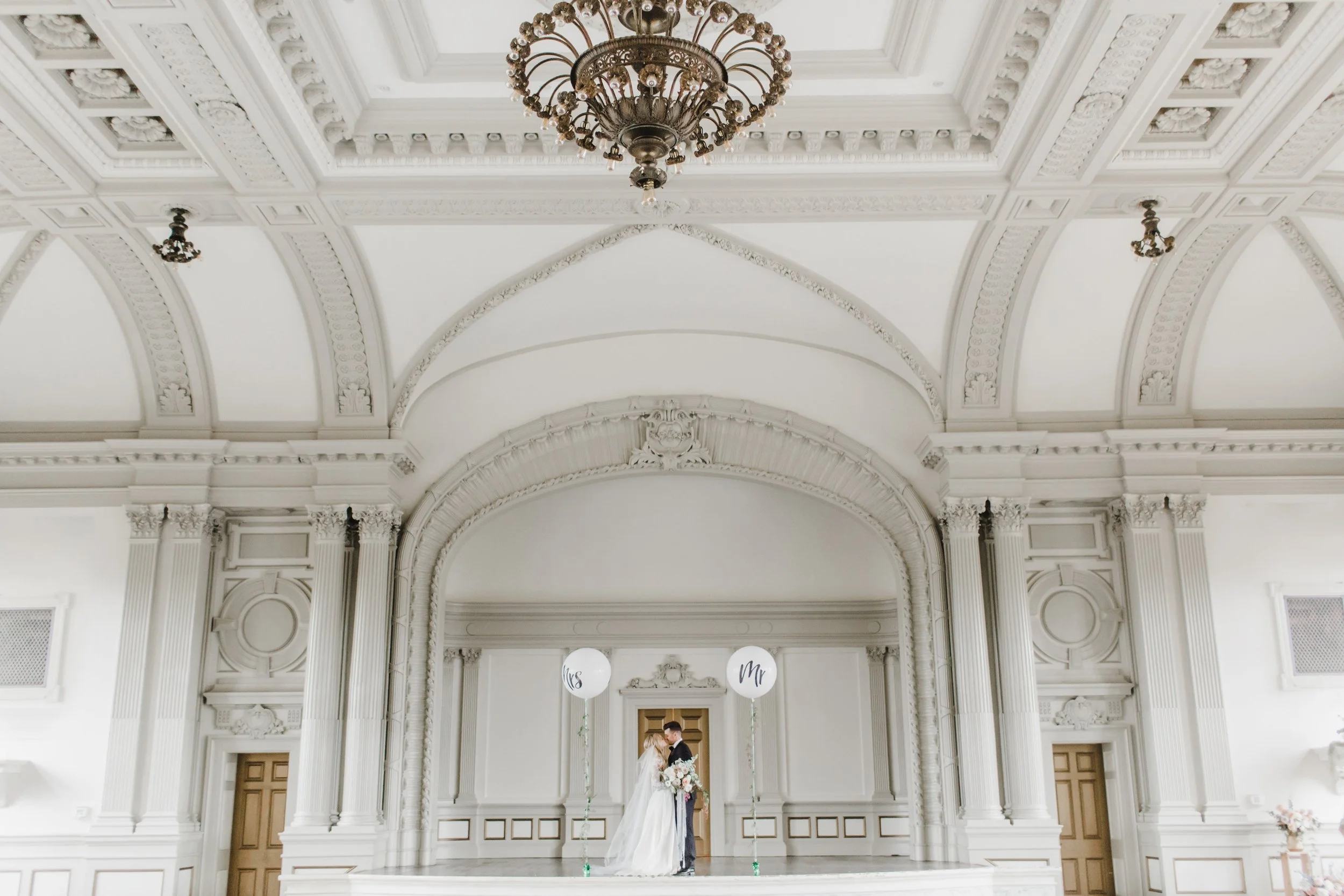 A bride and groom standing on a stage inside a grand, white, ornate hall during their wedding ceremony, with balloons that say "Mrs" and "Mr".