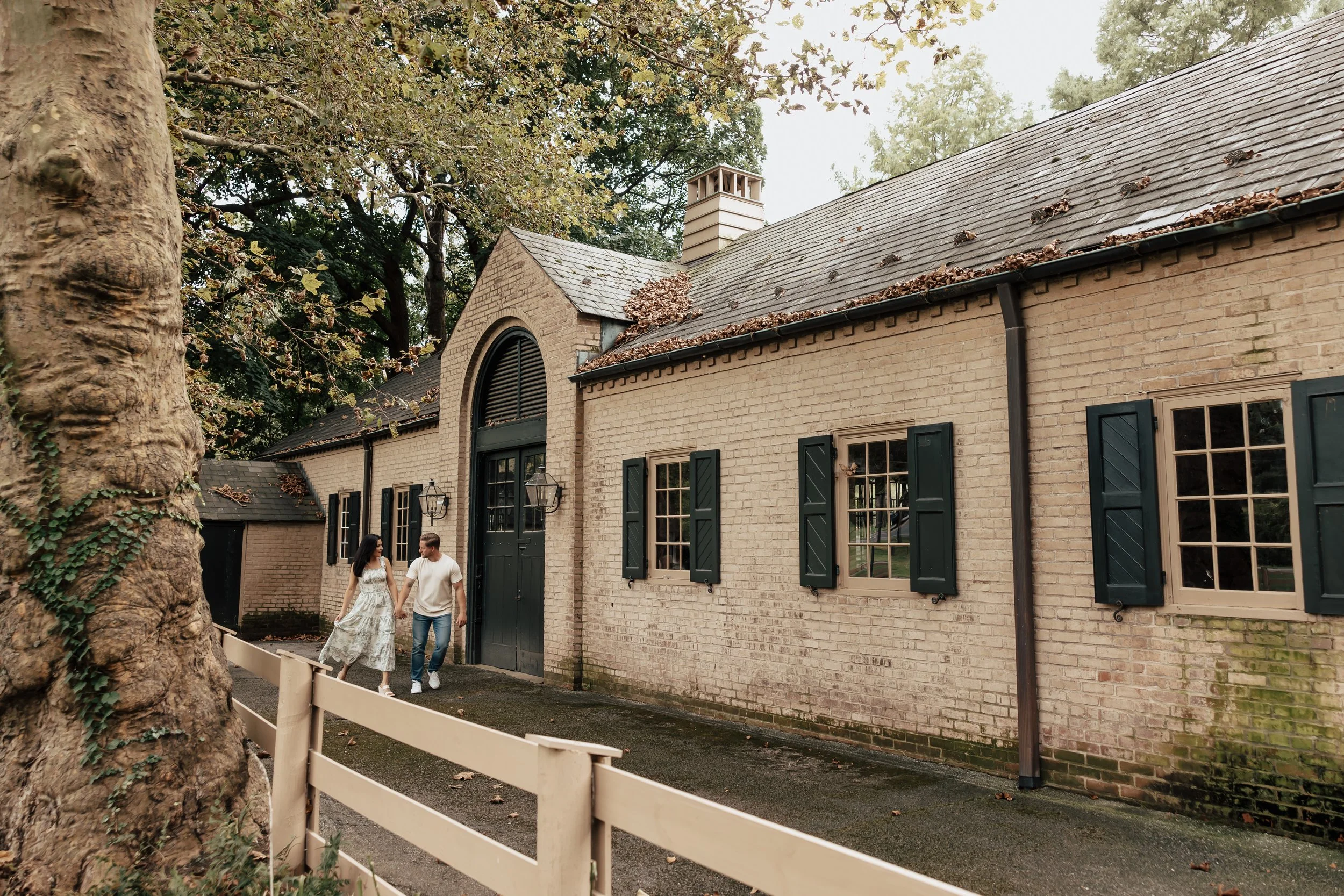 A couple walking hand in hand beside a rustic brick house with black window shutters, surrounded by trees with falling leaves.