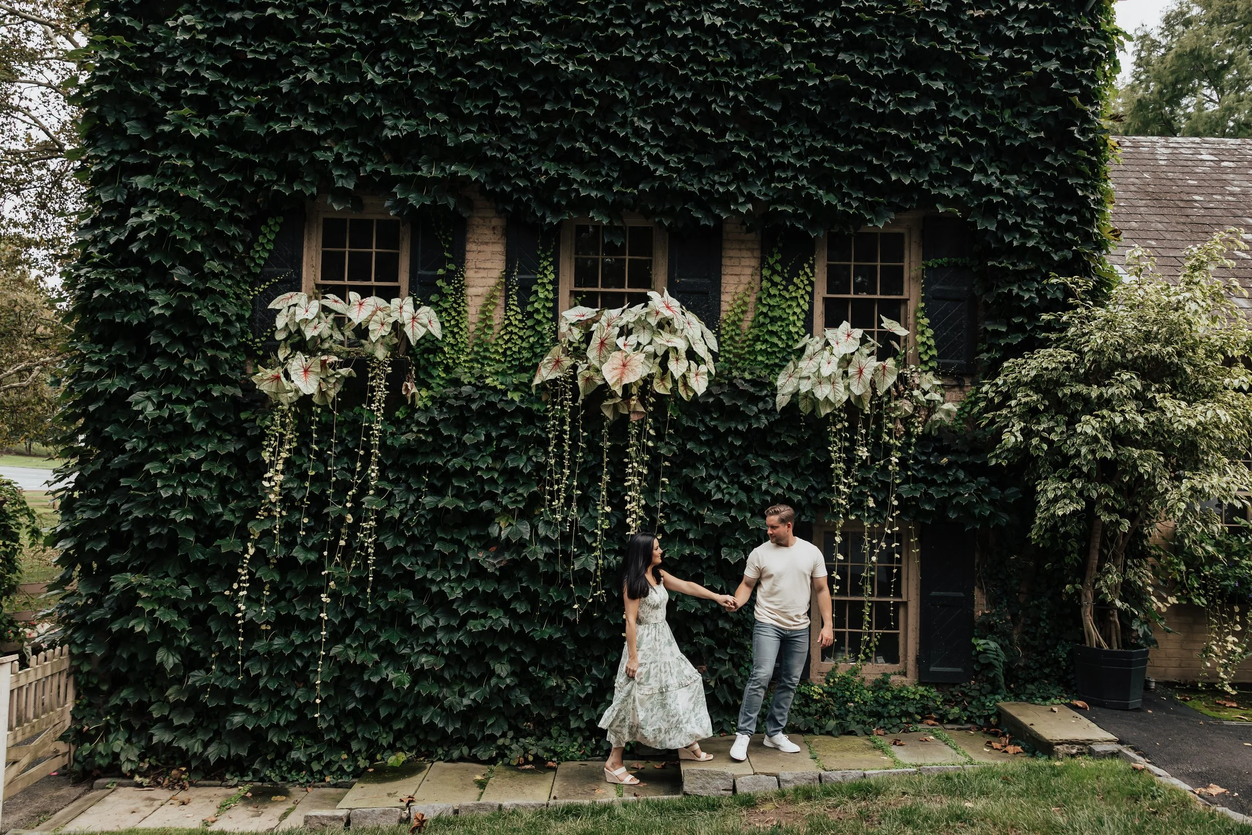 A couple holding hands while walking in front of a plant-covered house with large climbing plants and ivy.
