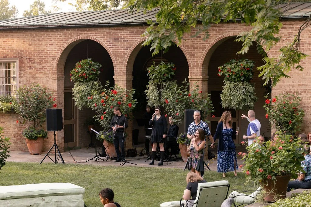 People gathered on a patio with a musical band performing, surrounded by large potted plants and hanging flower baskets, in front of a brick building with arches.