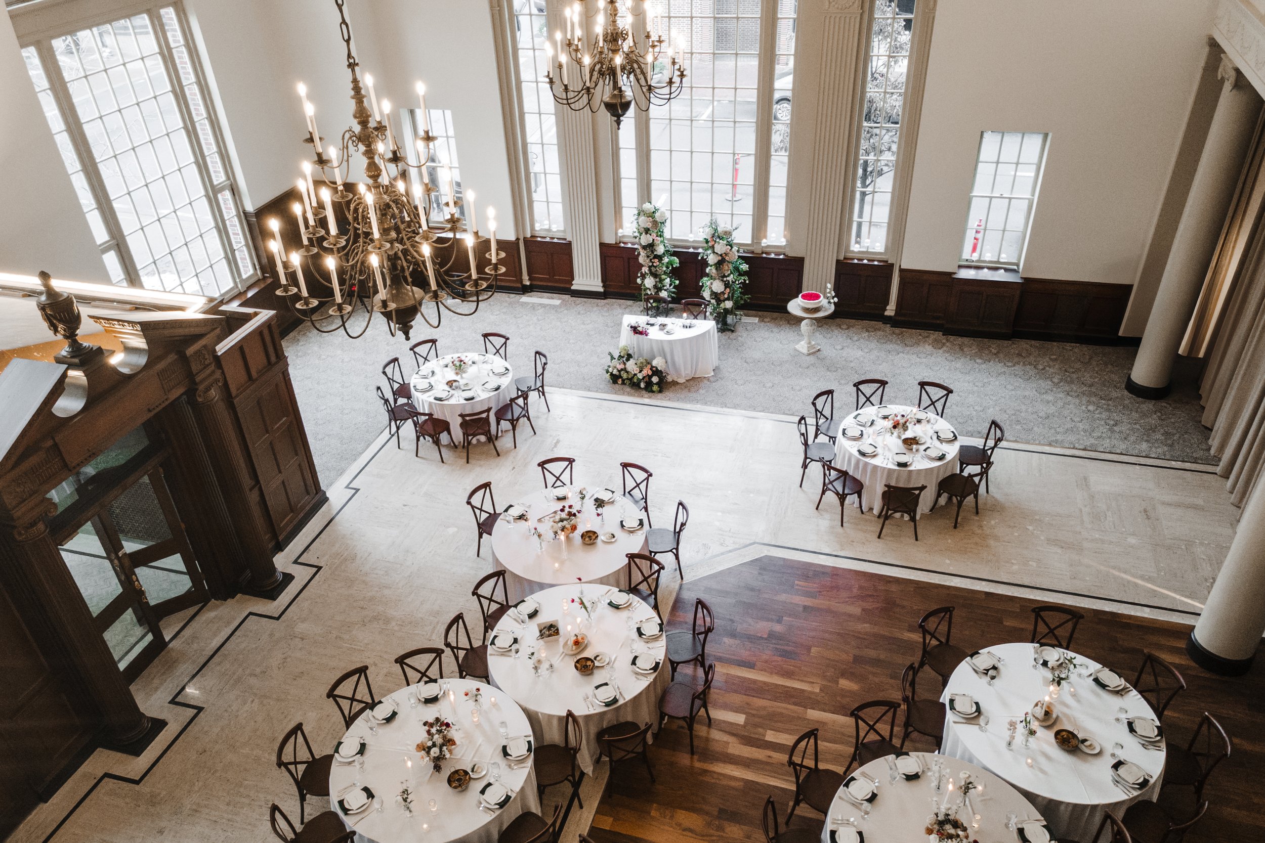 Elegant banquet hall decorated for a wedding or event with round tables, floral centerpieces, a chandelier, and large windows.