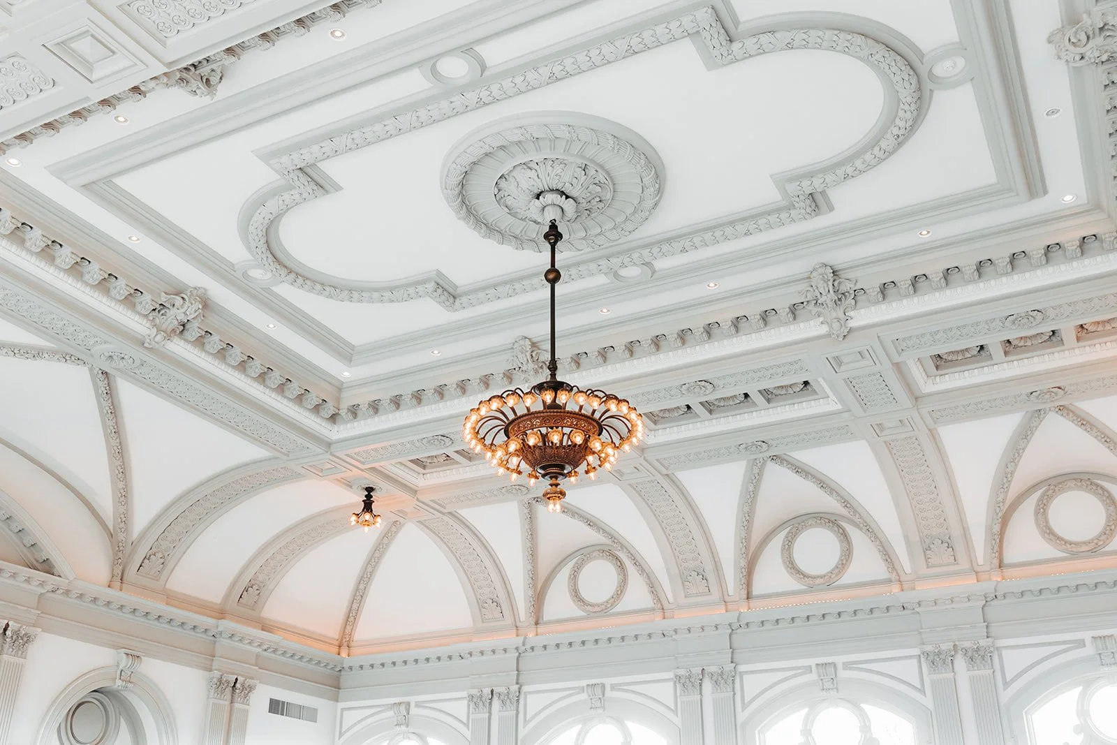 Elegant white ceiling with intricate moldings and patterns, central chandelier with multiple lights, and large arched windows allowing natural light.