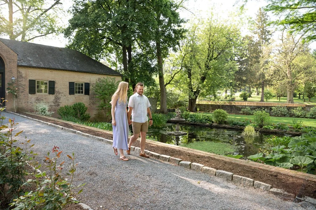 A couple walking along a gravel path beside a pond in a lush garden with trees and green foliage, a brick house with black shutters in the background.
