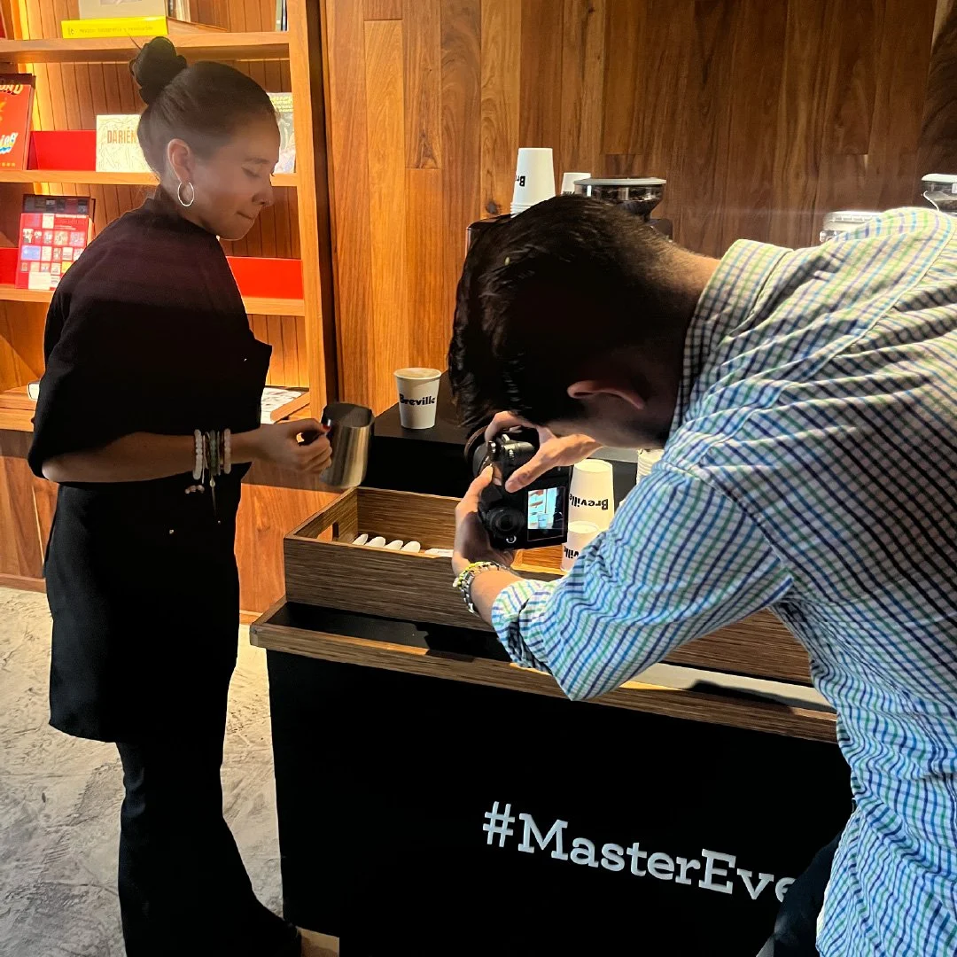 Una mujer sirviendo una bebida en una cafetería mientras un hombre toma una fotografía de ella. breville activacion pdv