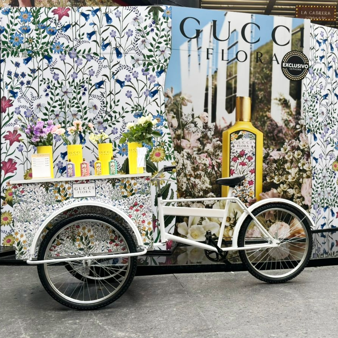 Carretilla decorada con flores, exhibiendo productos de Gucci Flora, junto a una bicicleta blanca de tres ruedas en un punto de exhibición con fondo floral y publicidad de Gucci Flora.