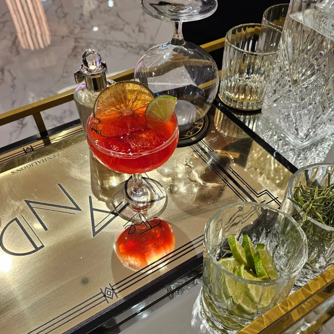 Cóctel rojo en copa de cóctel, decorado con rodaja de limón seca y rodaja de limón verde, junto a varias bebidas de cristal y adornos en bandeja dorada sobre mesa de mármol.