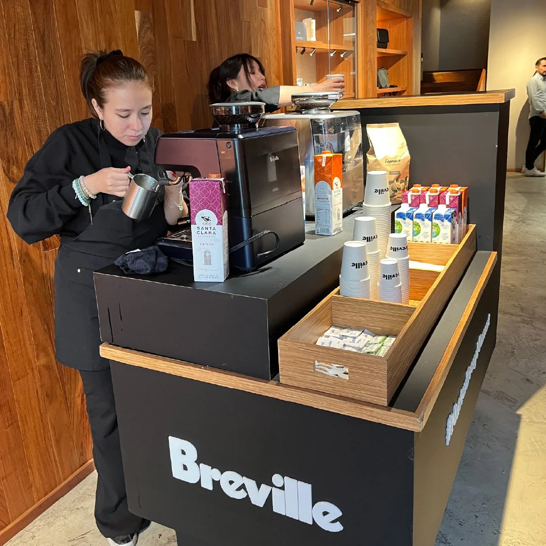 breville activacion. Una joven sirviendo café en una máquina de café en una cafetería llamada Breville mas bebidas y vasos apilados en el mostrador.