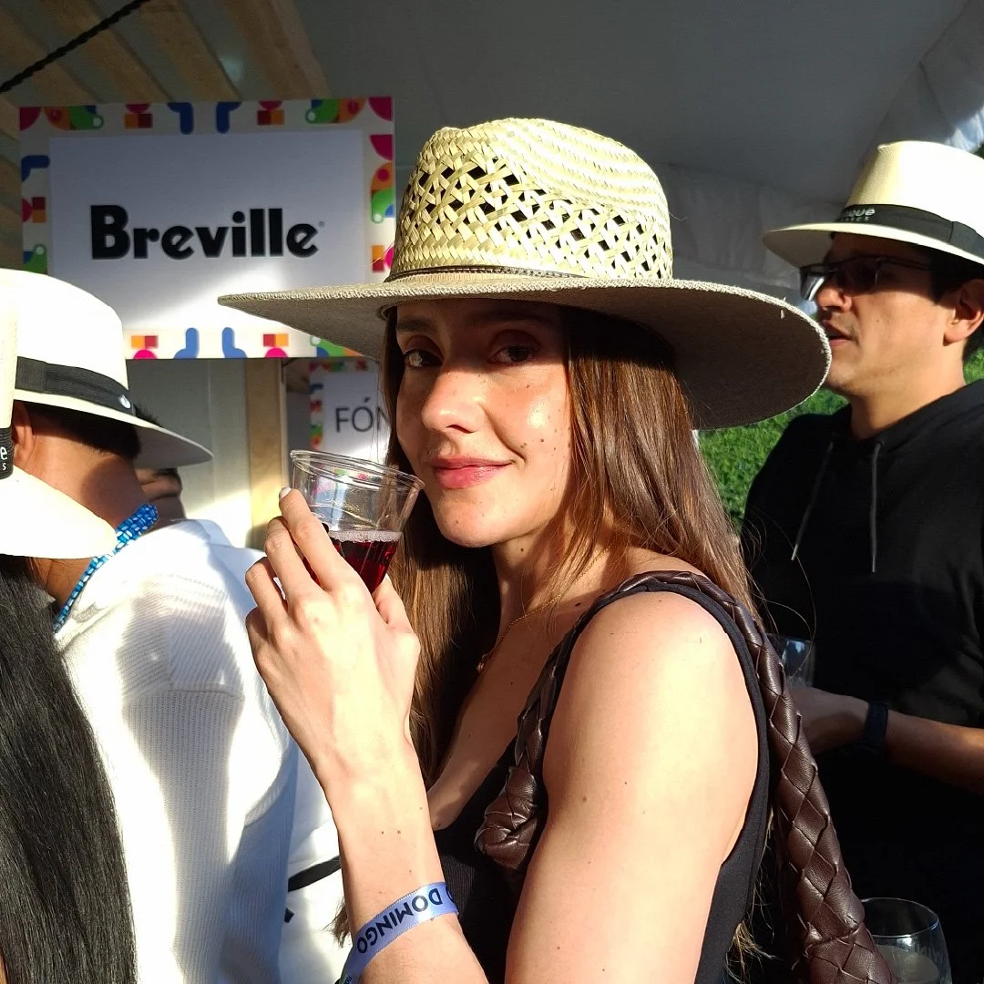 Mujer con sombrero de paja y vestido negro, sosteniendo una copa de vino tinto en un evento con otras personas, algunos con sombrero similar, y logotipo de Breville en el fondo.