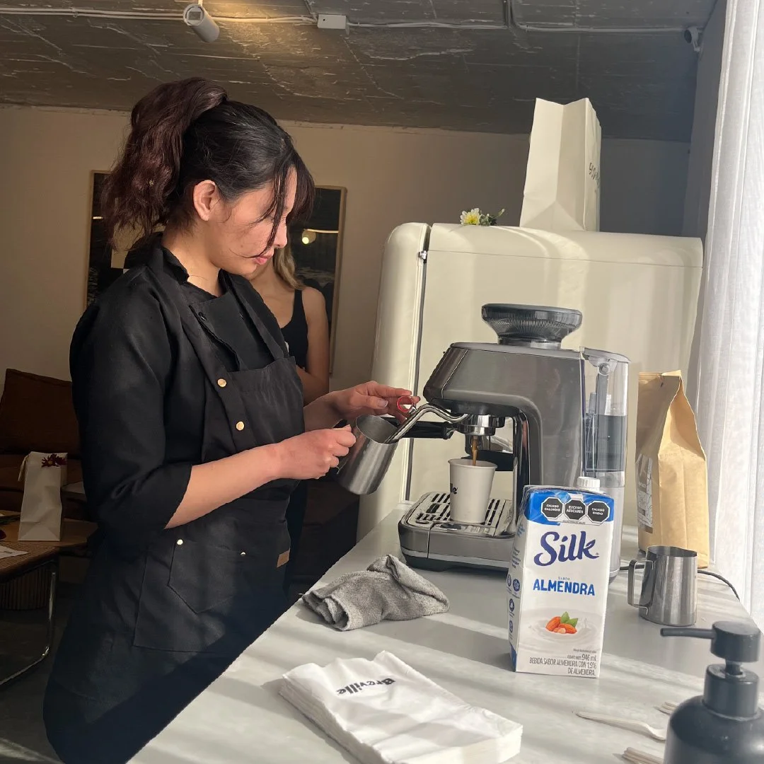 Una mujer sirviendo café con una máquina de espresso en un mostrador de cafetería, con leche de almendra en un envase al frente.