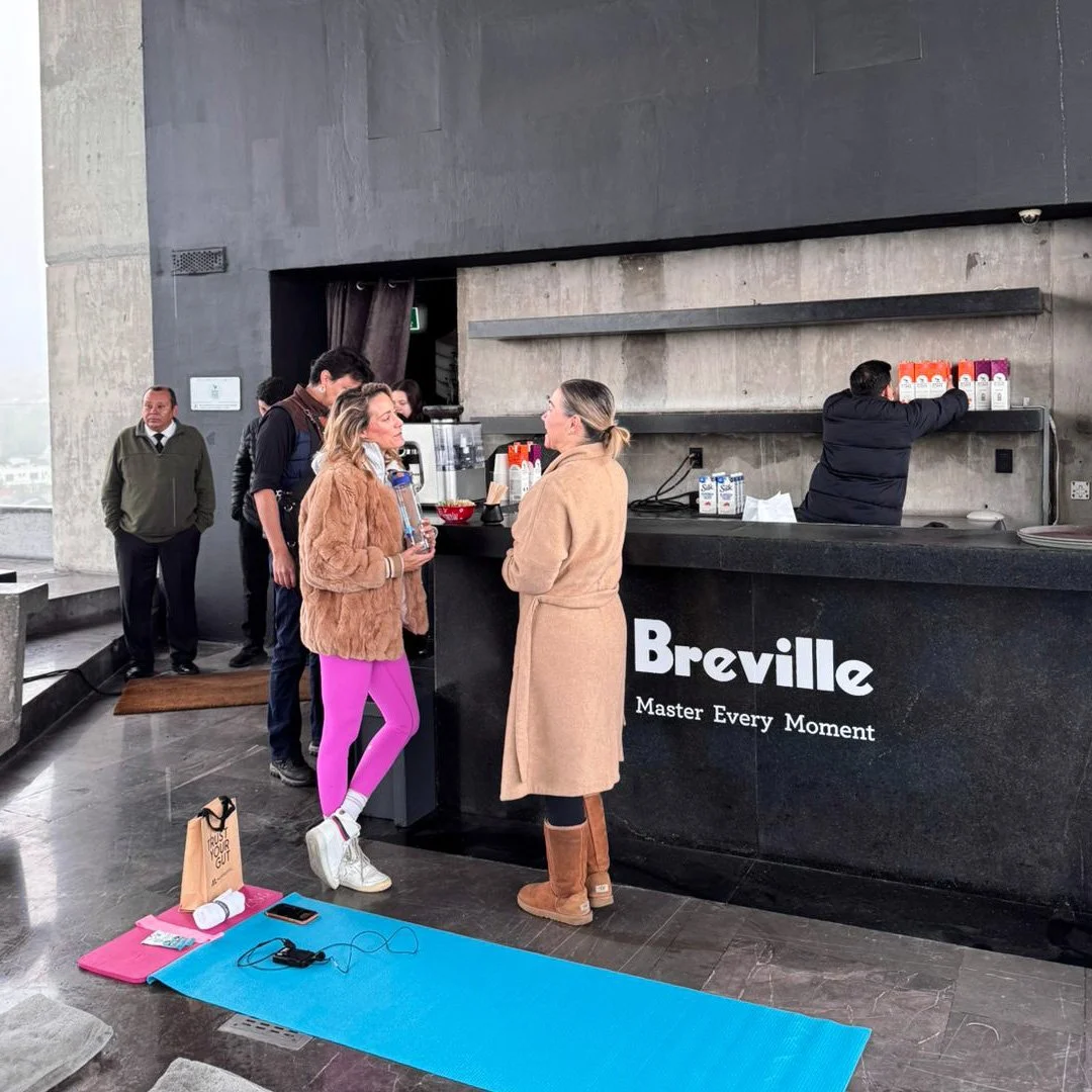 Personas en una cafetería en una ciudad, con un mostrador de bebidas, y personas conversando y haciendo fila para ordenar, en un entorno moderno con paredes de concreto.
