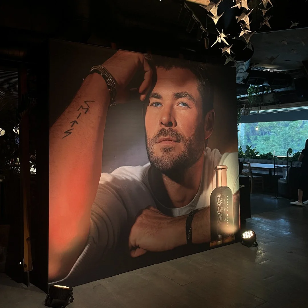 Cartel grande con la imagen de un hombre de cabello oscuro, barba, usando una camiseta blanca, que mira pensativo con el brazo apoyado y una frase escrita en el antebrazo, en un entorno interior con plantas y luz natural entrando por la ventana.