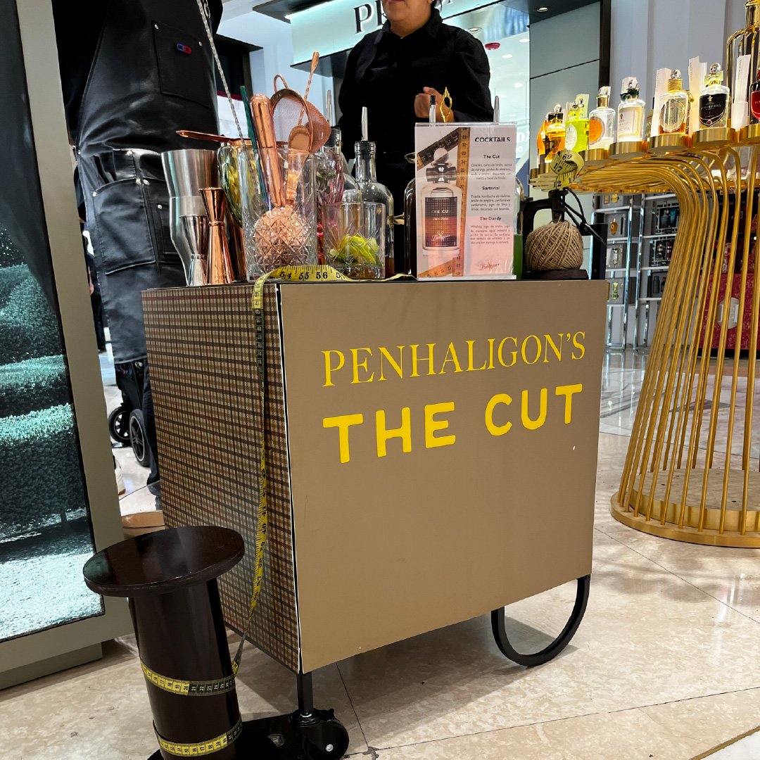 Carrito de bebidas en una tienda, con el letrero que dice 'Penhaligon's The Cut', mostrando frascos de perfume y utensilios de bar. activacion pdv