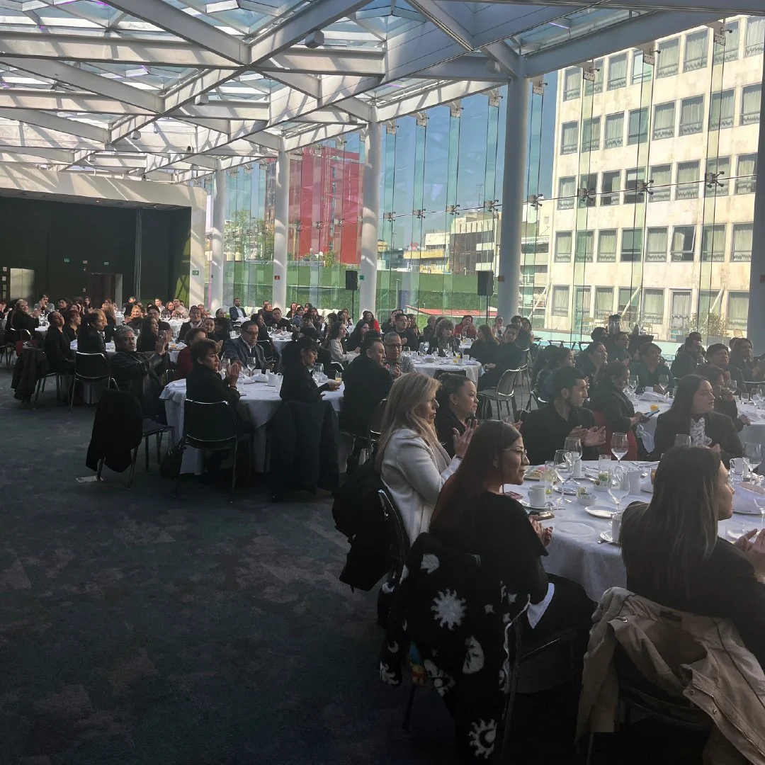 Imagen de una gran reunión o evento en un salón con paredes de cristal, con muchas personas sentadas en mesas cubiertas con manteles blancos, en un ambiente moderno y bien iluminado