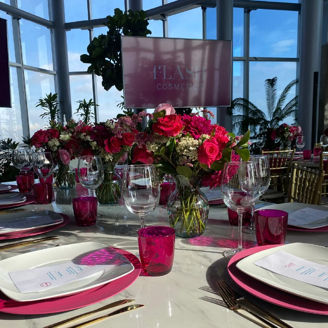 Mesa decorada con flores rosas y blancas en jarrones de vidrio, copas de vino, platos blancos con manteles rosados, vasos rosas, y un cartel en la pantalla que dice 'FLASH COSMETICS'.