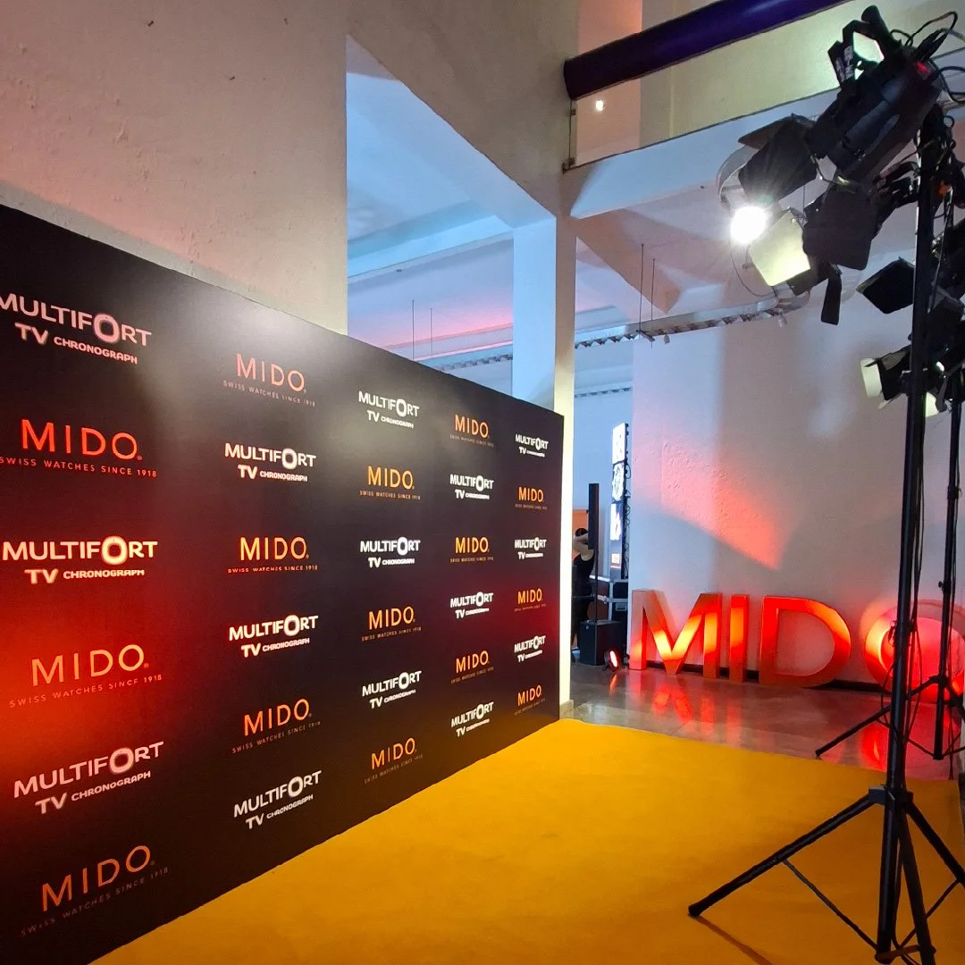 Logo wall de la marca MIDO en un evento con iluminación y fondo de escenario con letras en neón rojo que dicen MIDO, y un piso de alfombra naranja.
