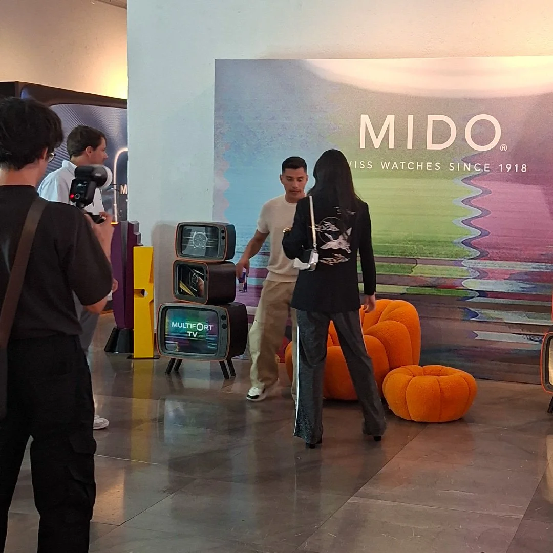Personas participando en una grabación o entrevista frente a un fondo con logotipo de MIDO, un reloj suizo, con muebles de color naranja y televisores vintage, en un entorno de interior con piso de mármol.