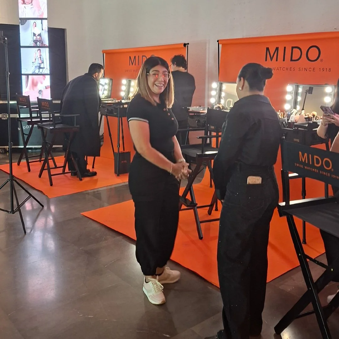 Dos mujeres conversando en un evento de maquillaje con un fondo naranja que dice MIDO, con estaciones de maquillaje y espejos con luces.
