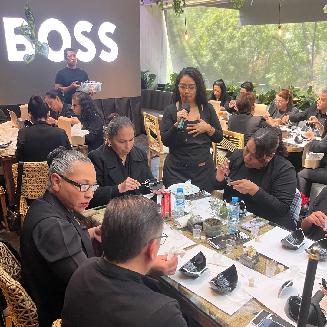 Mujeres y hombres en una reunión o comida empresarial en un restaurante con decoración moderna y muchas plantas, algunas personas revisando objetos, una mujer con gafas sosteniendo un micrófono, y un hombre en el escenario con la palabra 'BOSS' en la