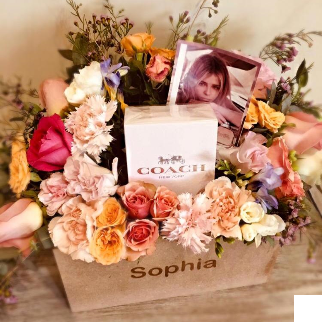 Arreglo de flores en una caja de madera con el nombre 'Sophia', con un collar de Coach, una foto de una mujer y una caja pequeña. influencer kit