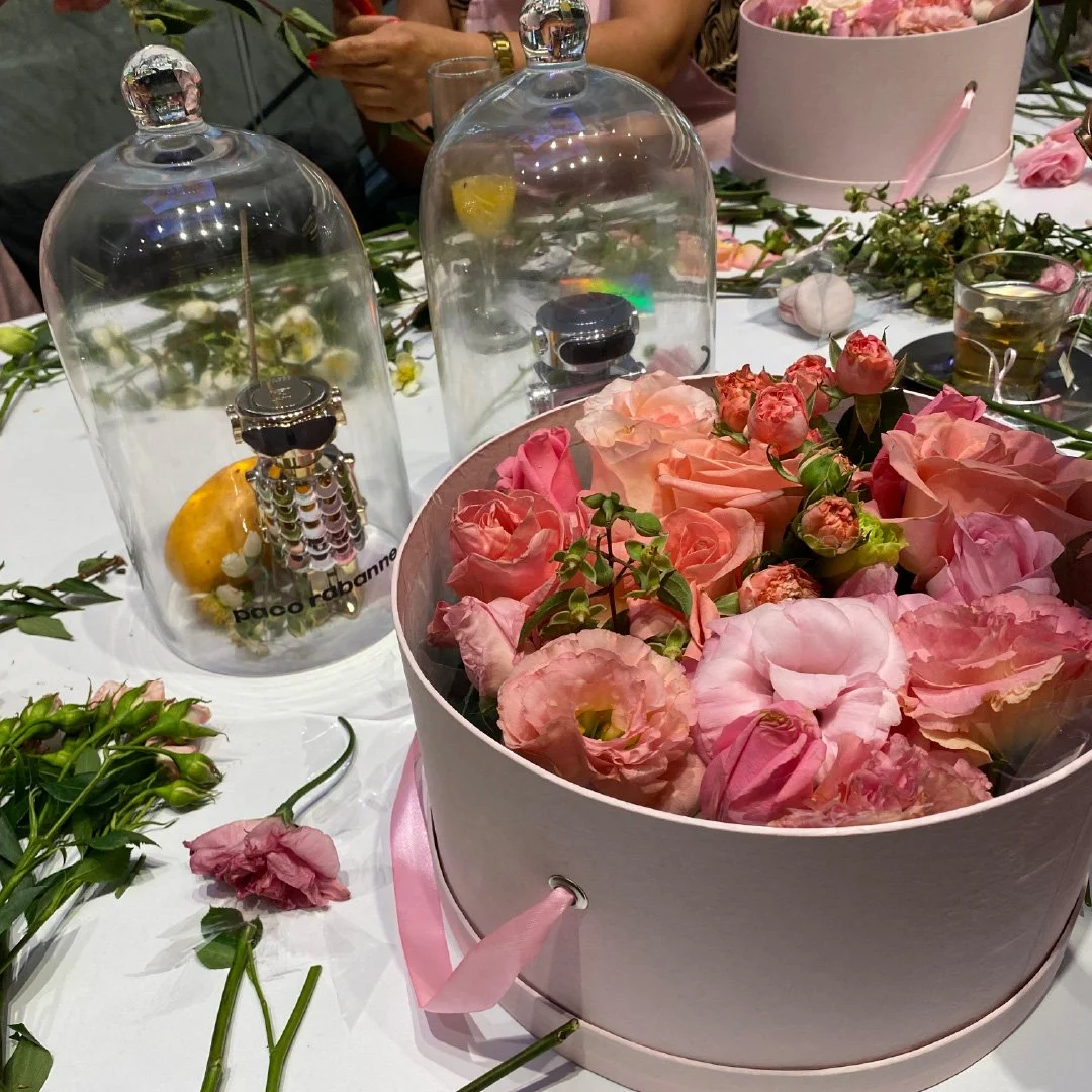 Macetero con flores rosas y pequeños racimos, varias flores y objetos decorativos en la mesa, personas en el fondo