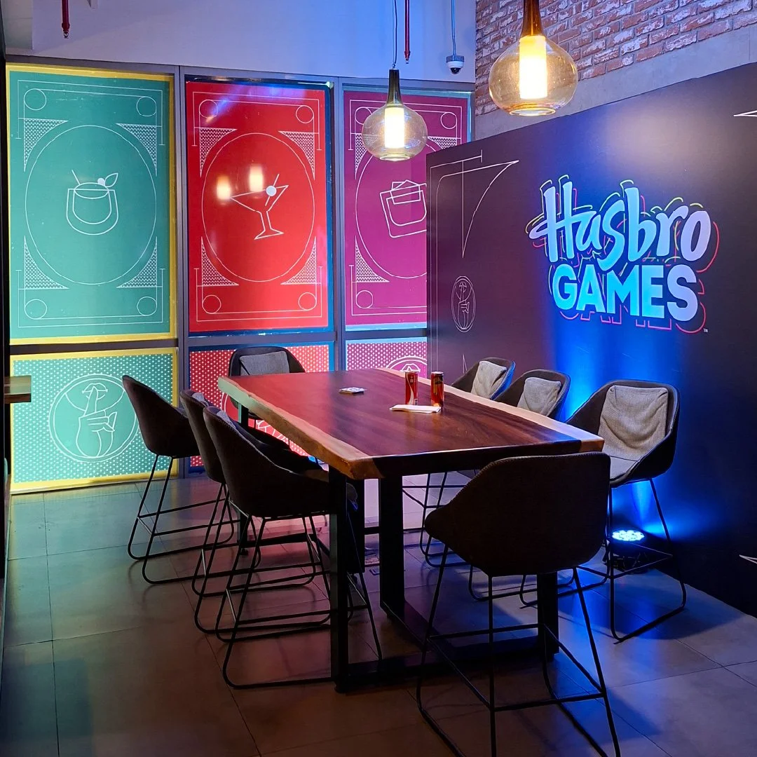 Sala de reuniones con cartel de "Hasbro Games" y paredes decoradas con ilustraciones de bebidas y objetos en colores vibrantes.