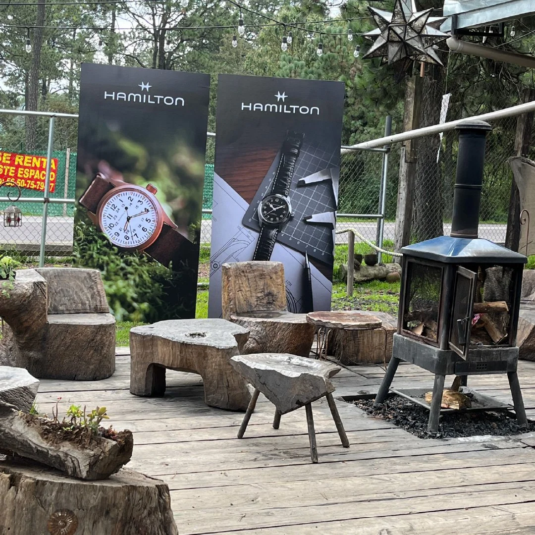 Anuncio de relojes Hamilton en un espacio al aire libre con sillas y una chimenea de madera