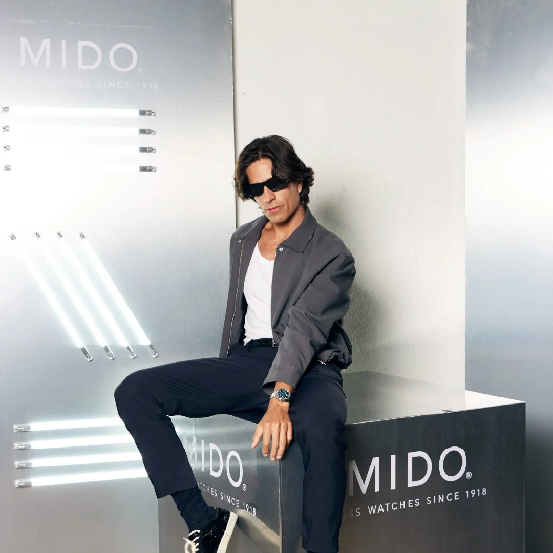 Hombre de cabello oscuro con gafas de sol, vistiendo una chaqueta gris, camiseta blanca y pantalones oscuros, sentado en un mueble con el logo de MIDO, una marca de relojes, en un entorno moderno con iluminación blanca.