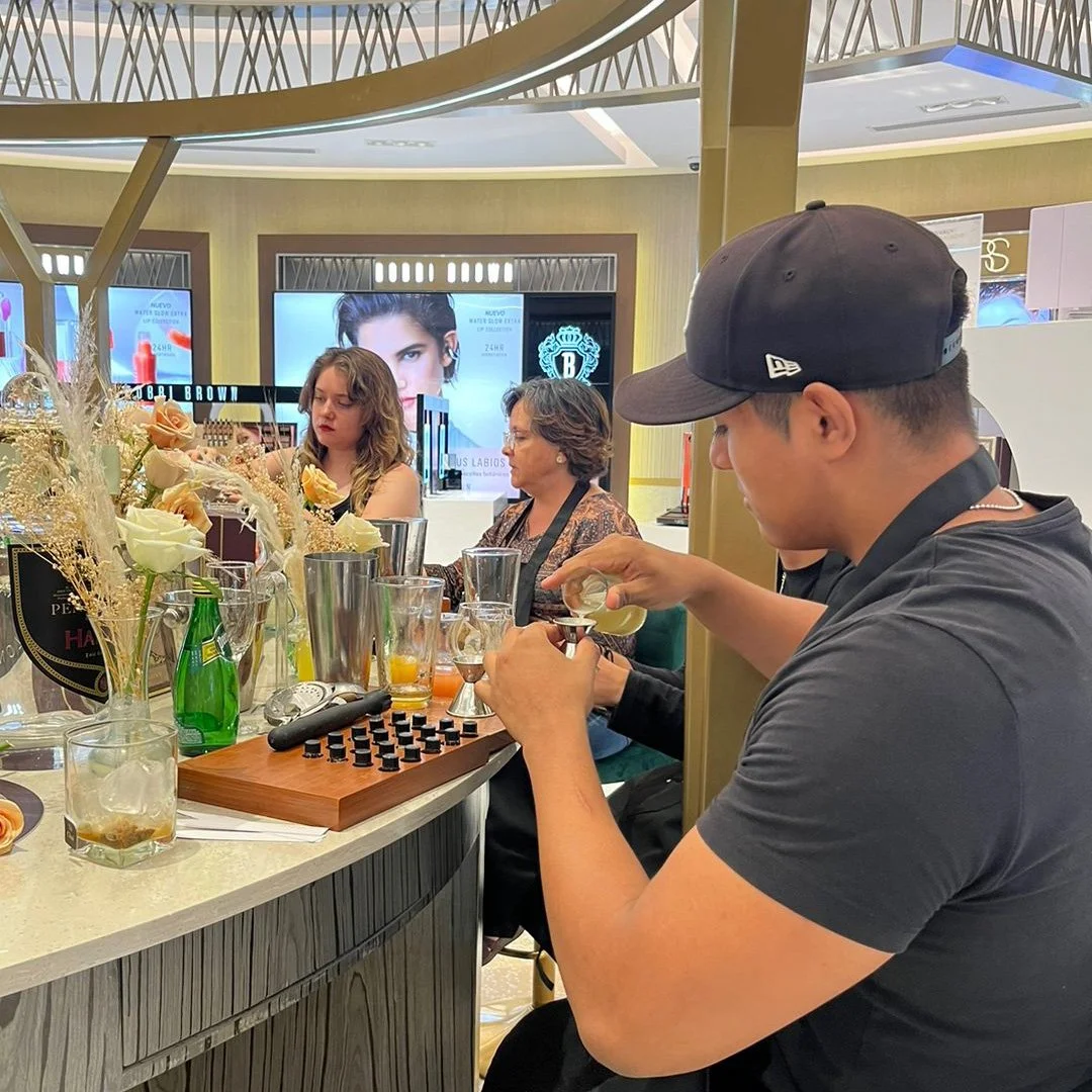 Un bartender preparando cócteles en una barra en un centro comercial, con varias personas observando y bebiendo.