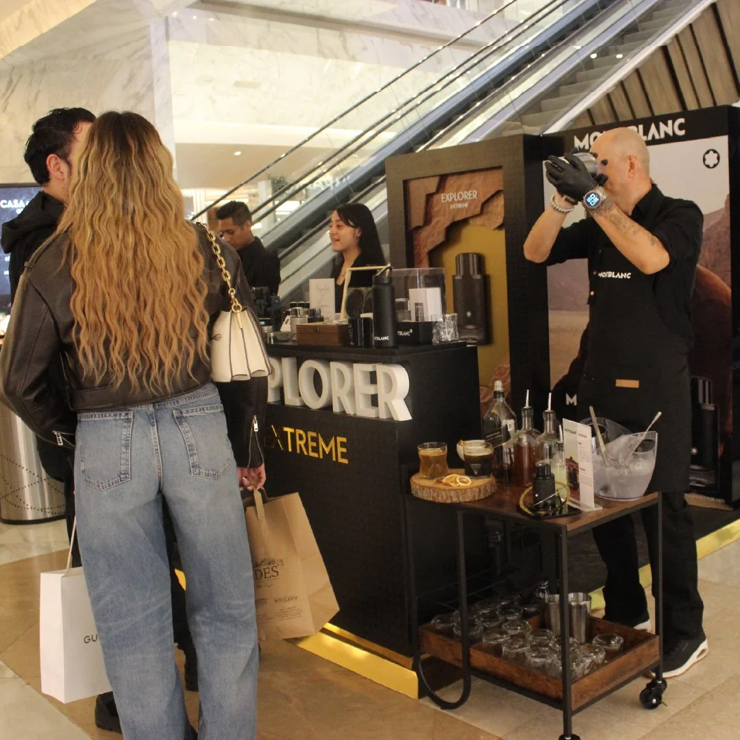 Personas en un puesto de degustación de bebidas en un centro comercial, atendido por un hombre con guantes negros y reloj inteligente, rodeados de productos y decoraciones de la marca Montblanc.