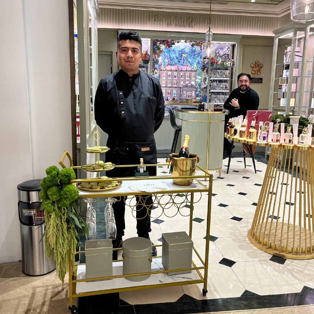 Hombre con uniforme negro en una mesa cobrando en una tienda de perfumes con decoraciones en tonos dorados y verdes, en el fondo una tienda con productos y un hombre sentado.