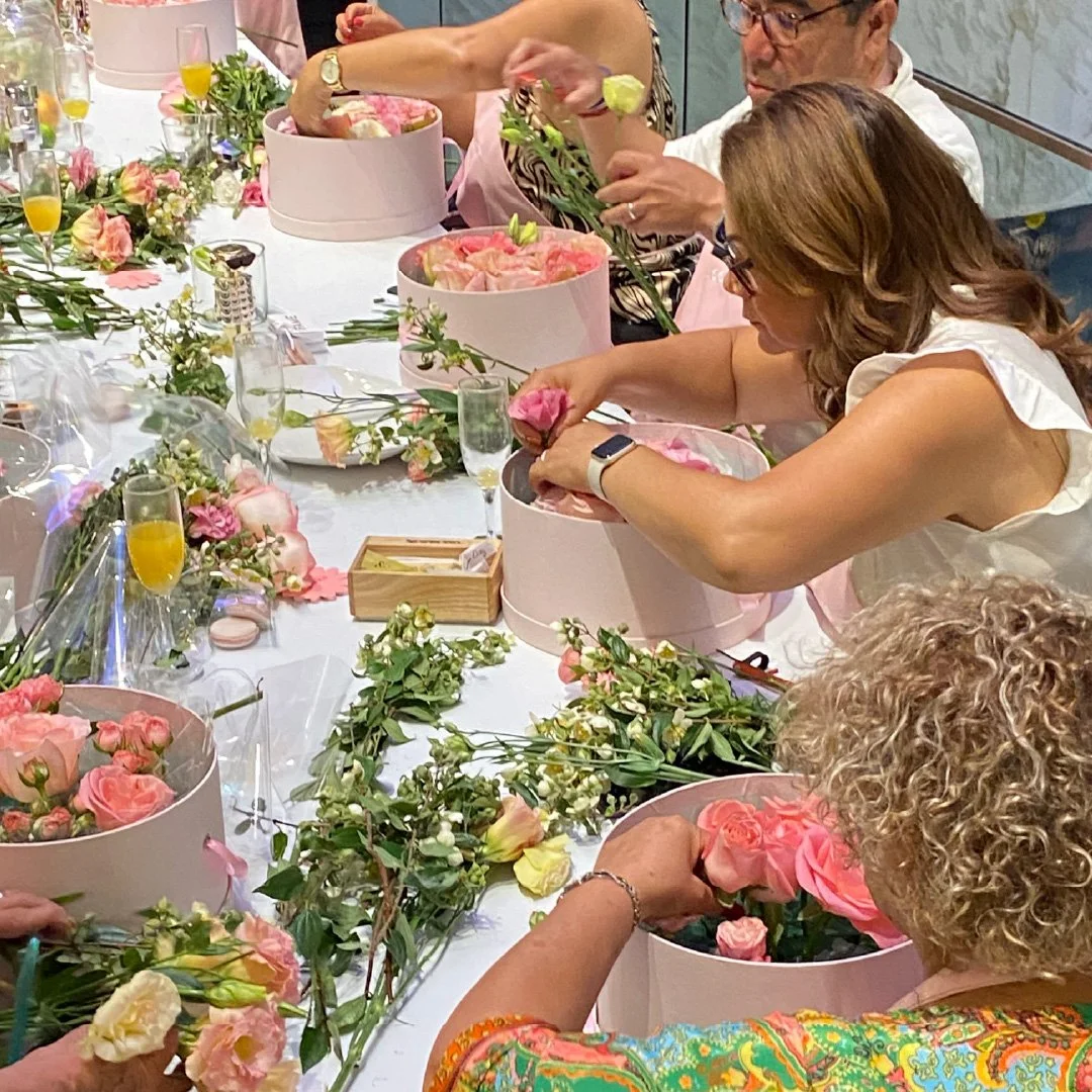 Varias personas decoran flores en una mesa con flores de color rosa, amarillo y blanco, y cajas redondas rosas. La mesa tiene varias copas con bebida y objetos decorativos. Las personas parecen estar preparando arreglos florales.