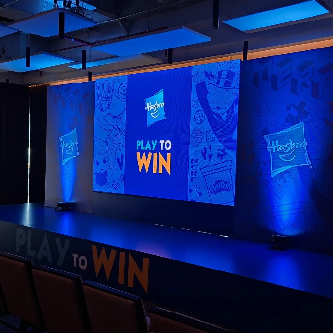 Pantalla digital con logo de Hasbro y el mensaje "Play to Win" en un escenario de presentación, decorado con ilustraciones de personajes y productos de la marca.