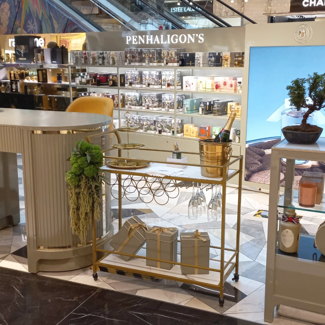 Display de productos de perfumes en una tienda, con una decoración elegante en tonos dorados y blancos, includes un carrito con pequeñas cajas y una botella de champagne en un cubo dorado.