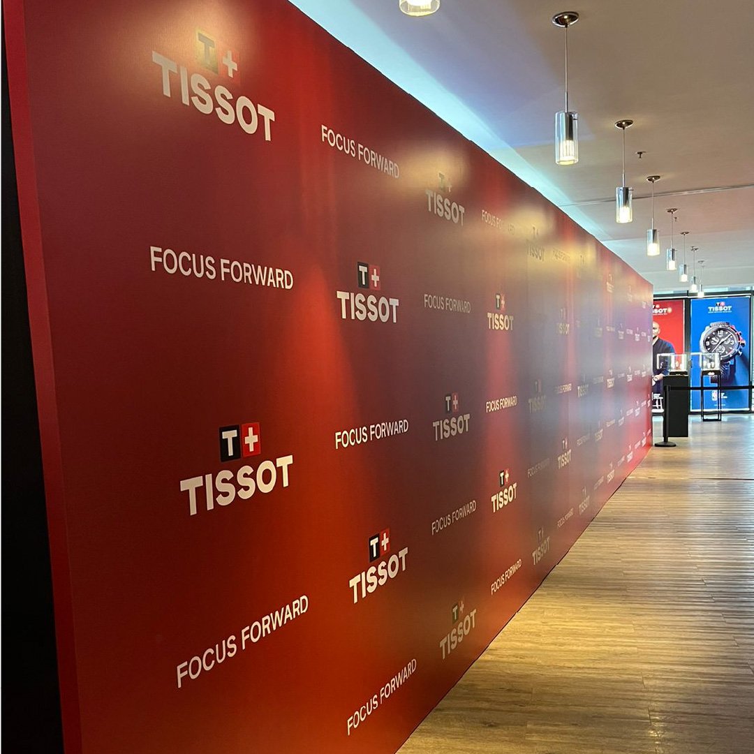 Pared roja con logotipos de Tissot y el lema 'Focus Forward' repetidos en un corredor de tienda o exhibición, con techo blanco y lámparas colgantes. Tissot Training mexico