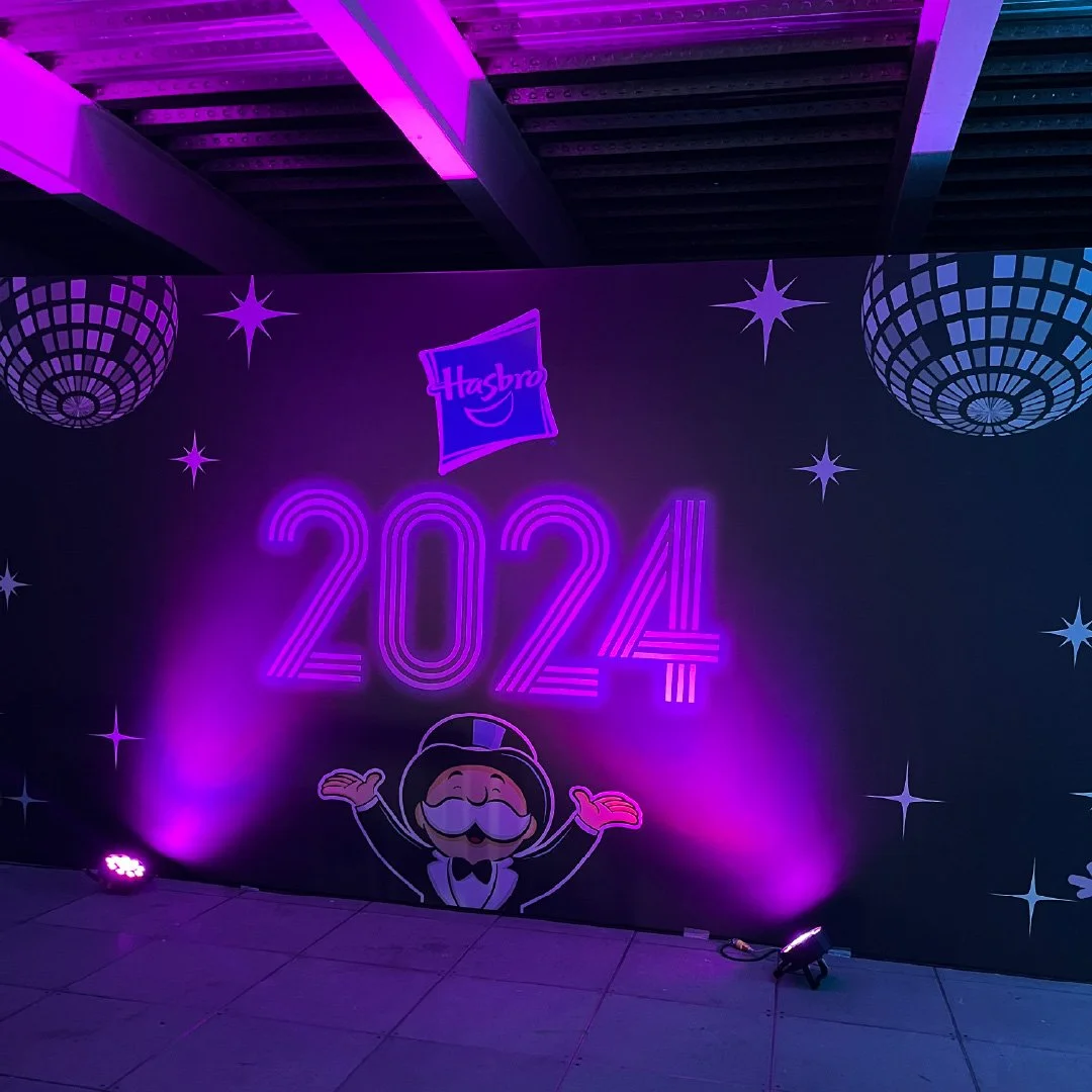Fondo decorativo con logotipo de Hasbro, años 2024 en luces de neón moradas, con personajes animados y estrellas, iluminación de escenario.