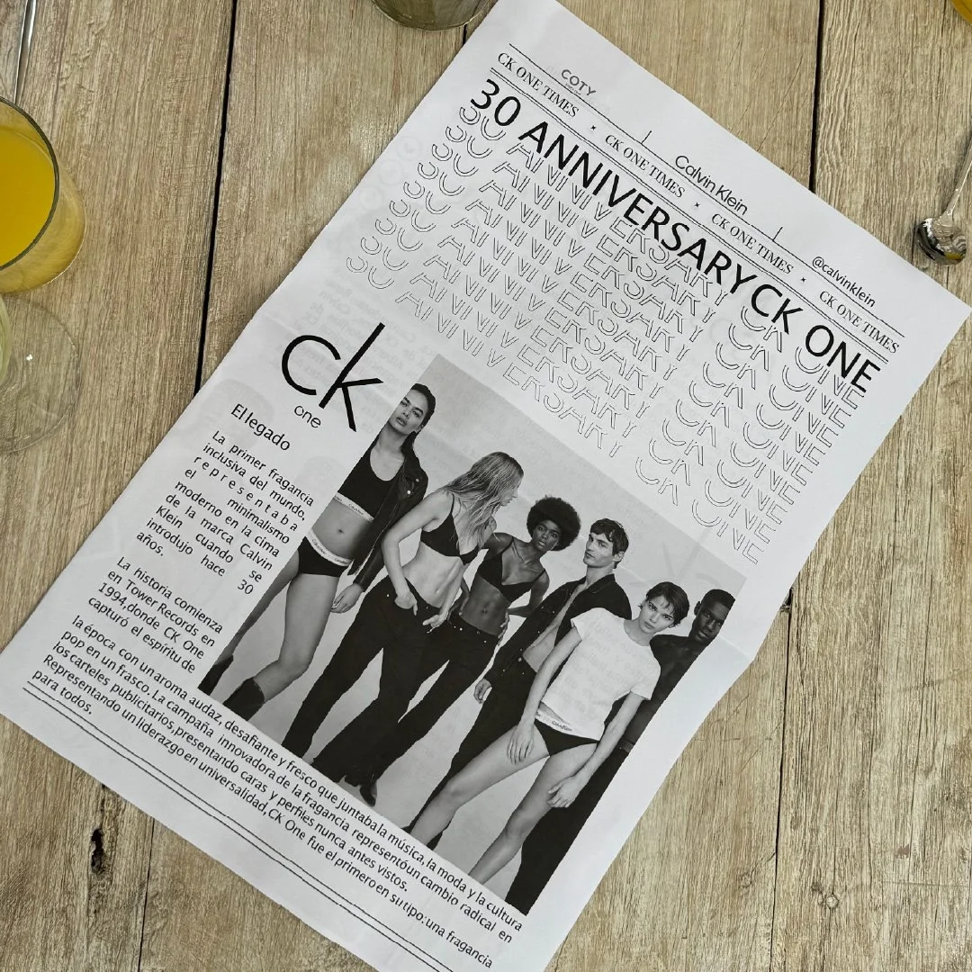 Página impresa con información en español sobre el 30 aniversario de CK One, con foto en blanco y negro de seis modelos jóvenes, cuatro mujeres y dos hombres, en ropa moderna y algo atlética, en un fondo blanco, sobre mesa de madera.