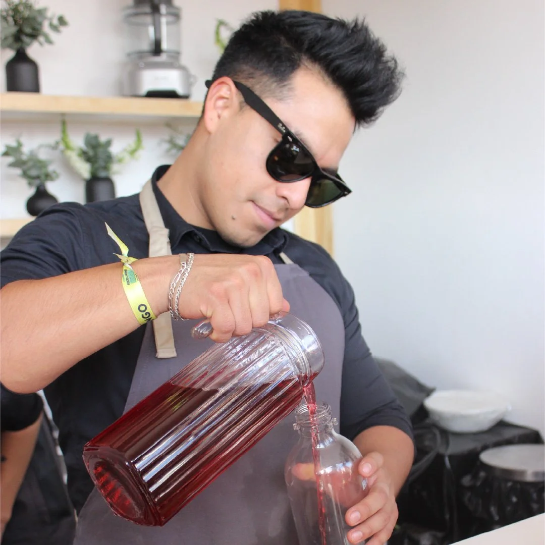 Un hombre joven con gafas negras vertiendo un líquido rojo en una botella de vidrio en un ambiente de cocina moderna.