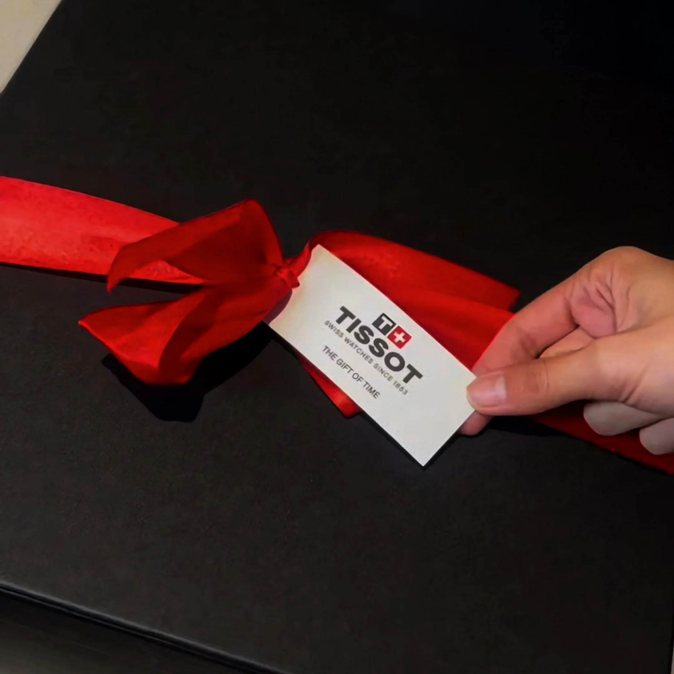 Mano sosteniendo una caja de regalo con cinta roja y una etiqueta que dice TISSOT, reloj suizo, y el eslogan "The gift of time".