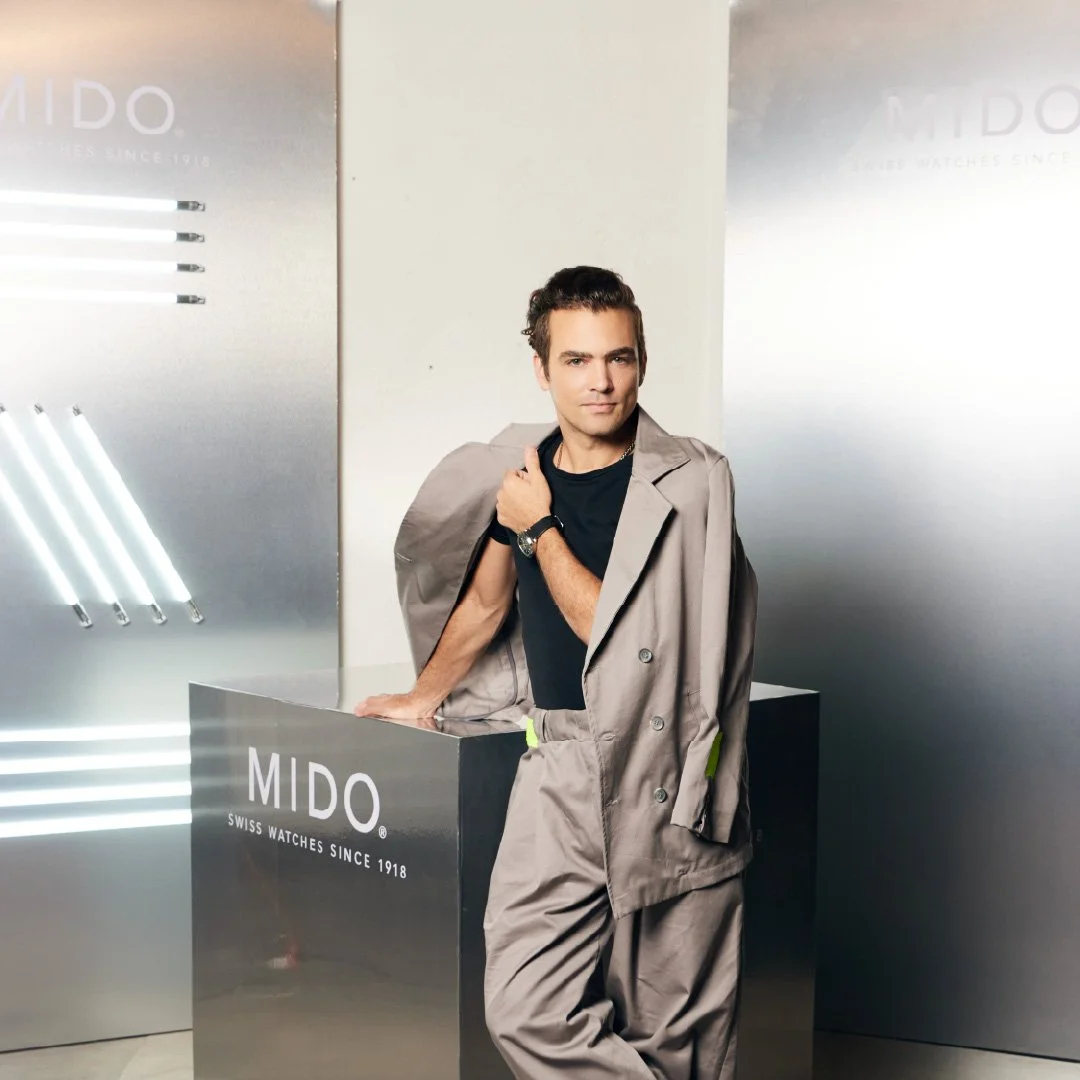 Un hombre posa junto a un mostrador con el logotipo MIDO, en un ambiente con decoraciones de acero y luces blancas. Está vestido con ropa en tonos gris y negro, con una chaqueta colgada sobre sus hombros.