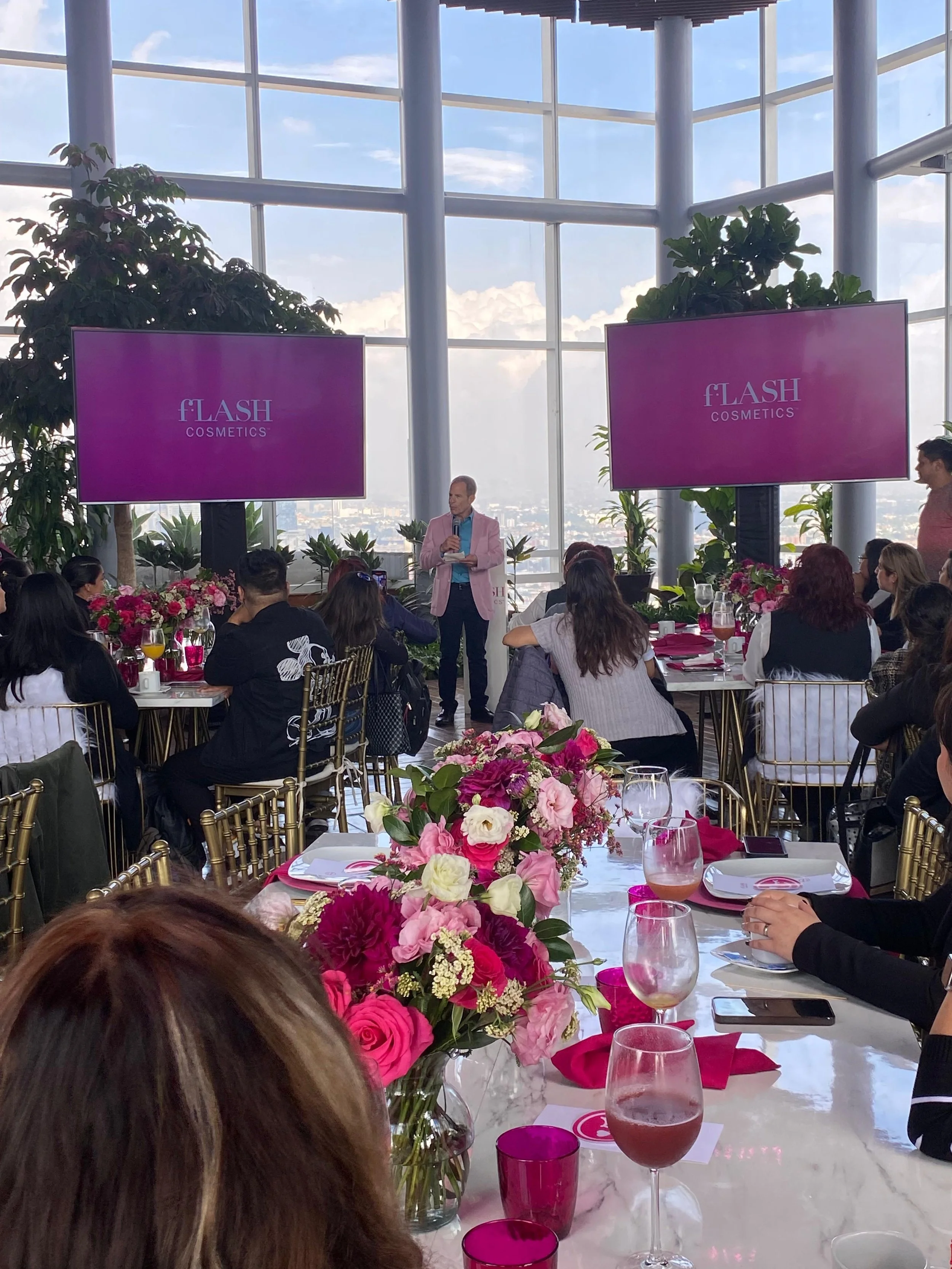 Conferencia en un lugar acristalado con muchas plantas y decoraciones florales en mesas, presentador de pie ante pantallas que dicen 'fLASH COSMETICS', y asistentes sentados observando.