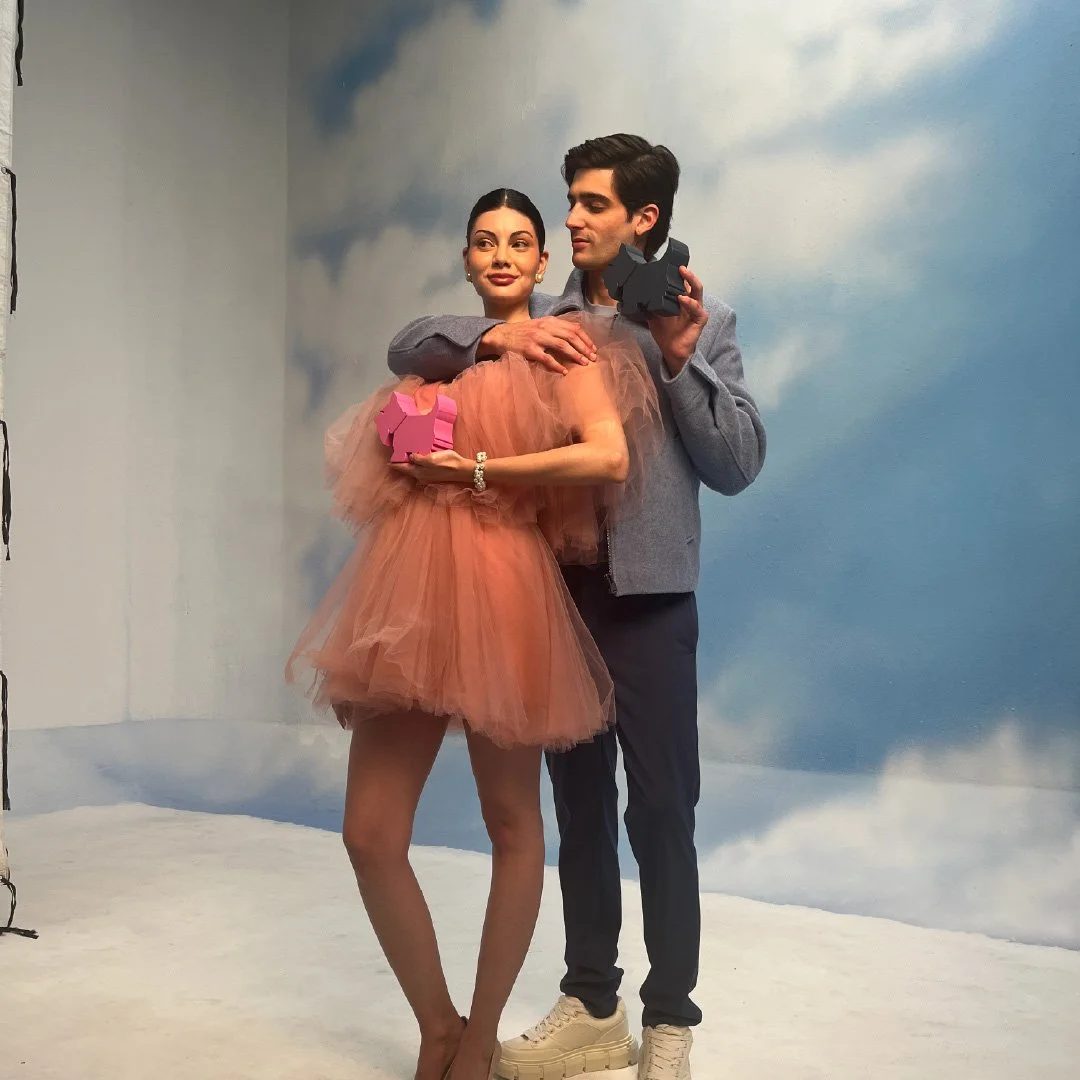 Un hombre y una mujer posando juntos en un escenario con fondo de cielo y nubes, ella con un vestido de tul naranja y sosteniendo una figura de unicornio rosa, él con un suéter gris y sosteniendo una cámara.