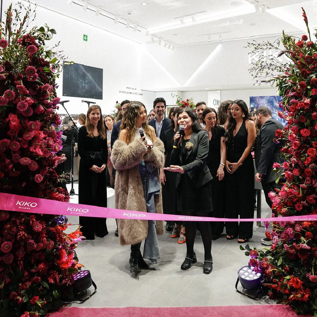 Evento en una galería de arte con muchas flores rojas y rosa, donde varias personas están participando en una inauguración, con dos mujeres en el centro hablando con micrófonos y un grupo de asistentes en el fondo.