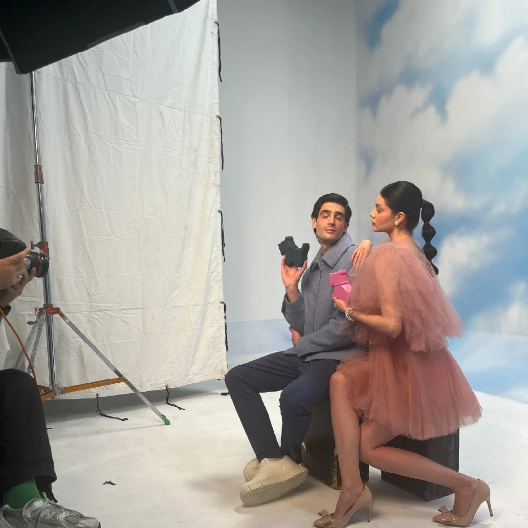 Dos personas posando en un estudio fotográfico con fondo de cielo y nubes. La mujer lleva un vestido de tul color durazno y tacones beige. El hombre veste casual con una chaqueta gris y pantalones oscuros, sosteniendo una cámara de mano.