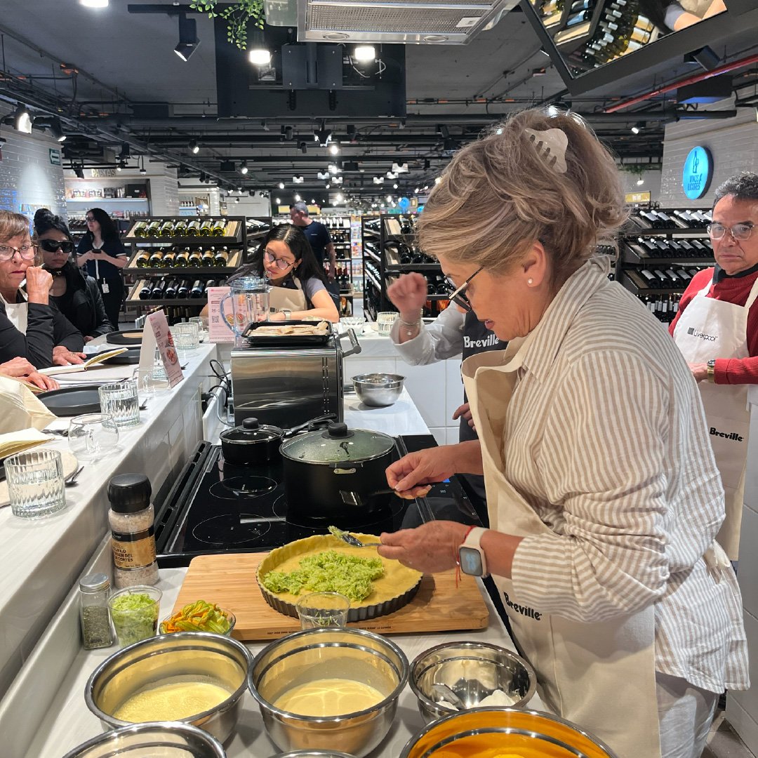 Mujer mayor cocinando en una cocina de un supermercado, con varias personas observando, preparando un plato con guacamole y otros ingredientes.